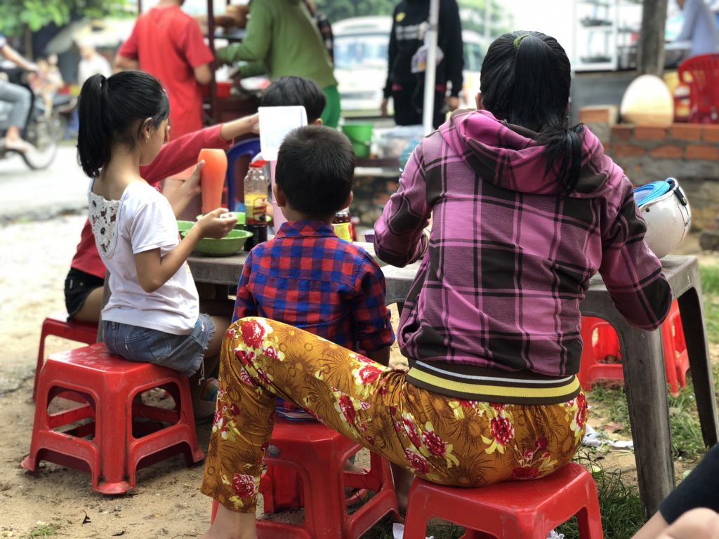 A mother and three children eating Vietnamese noodle dishes at a food stall